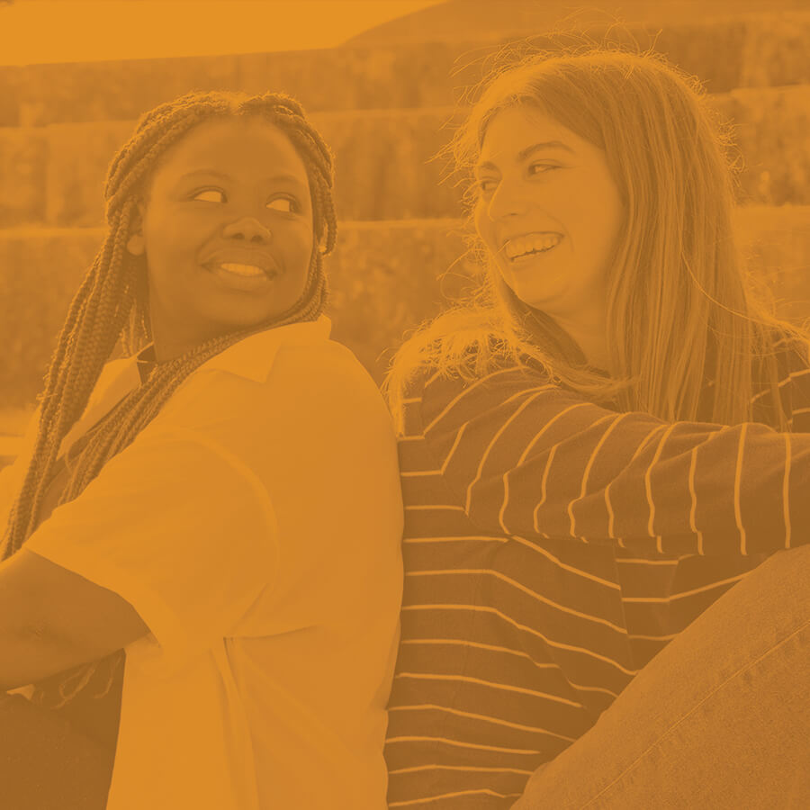 Two students smile at each other whilst sitting back-to-back on the bluff of Lake Michigan.