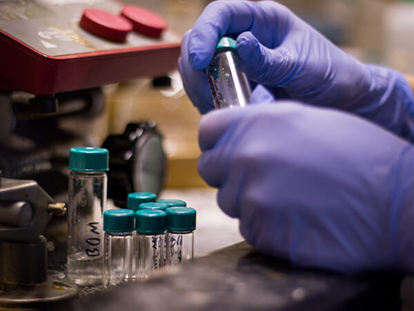 Student wearing blue lab gloves handling a small vial at a laboratory workbench.