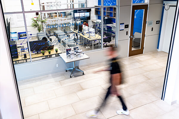 A view through the glass exterior of the modern AI & Quantum Innovation Lab at Concordia University Wisconsin, showing a bright workspace with computers and a blurred person walking by.