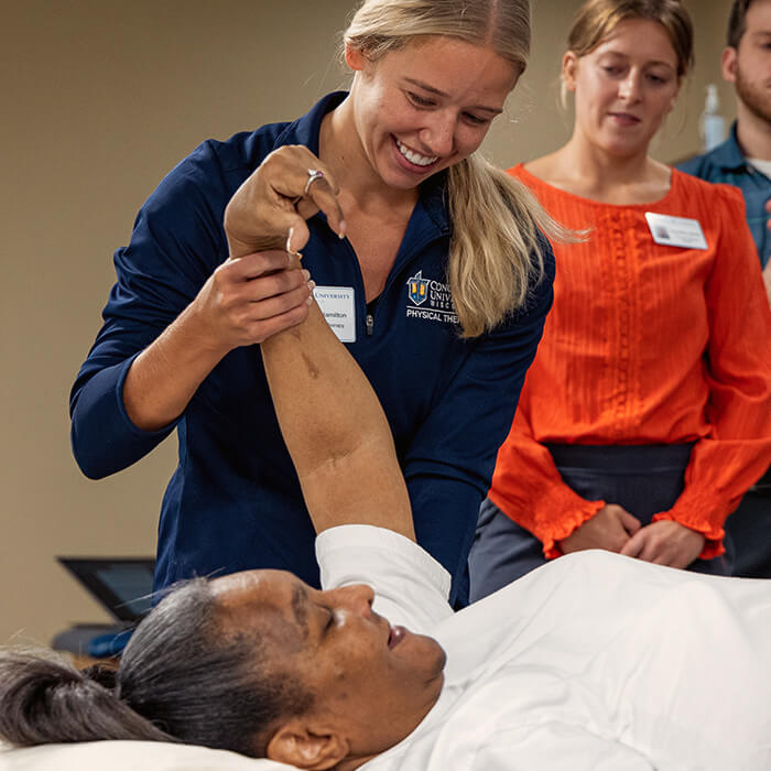 Physical therapy clinician assisting a patient with arm mobility while students observe in an interprofessional clinic setting.