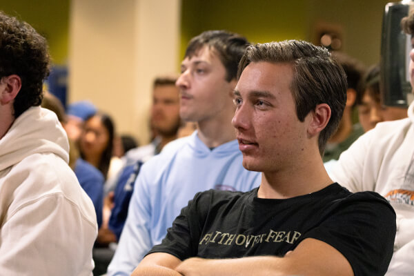A group of attendees at the Concordia National Security and Space Center's 'Luxembourg in Space' event, with a man in the foreground wearing a black 'FAITH OVER FEAR' T-shirt and several casually dressed students sitting behind him in a crowded indoor setting.